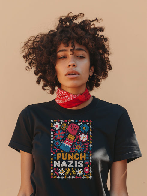 Black t-shirt featuring an embroidered-style design with the phrase "PUNCH NAZIS" in bold, colorful lettering. Above the text, a raised red fist is illustrated, symbolizing resistance, and is surrounded by a vibrant folk-art-inspired floral border with bright flowers, leaves, and geometric patterns in red, yellow, blue, and white.
